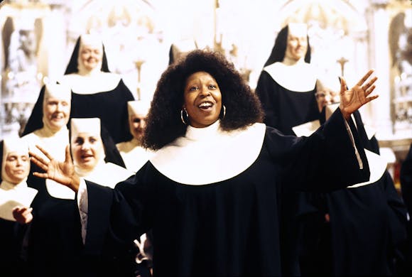 A woman in a habit sings with her arms outstretched in front of a choir of nuns.