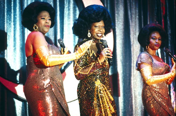 Three women with bouffant hairdos in sequened dresses sing into microphones on stage.