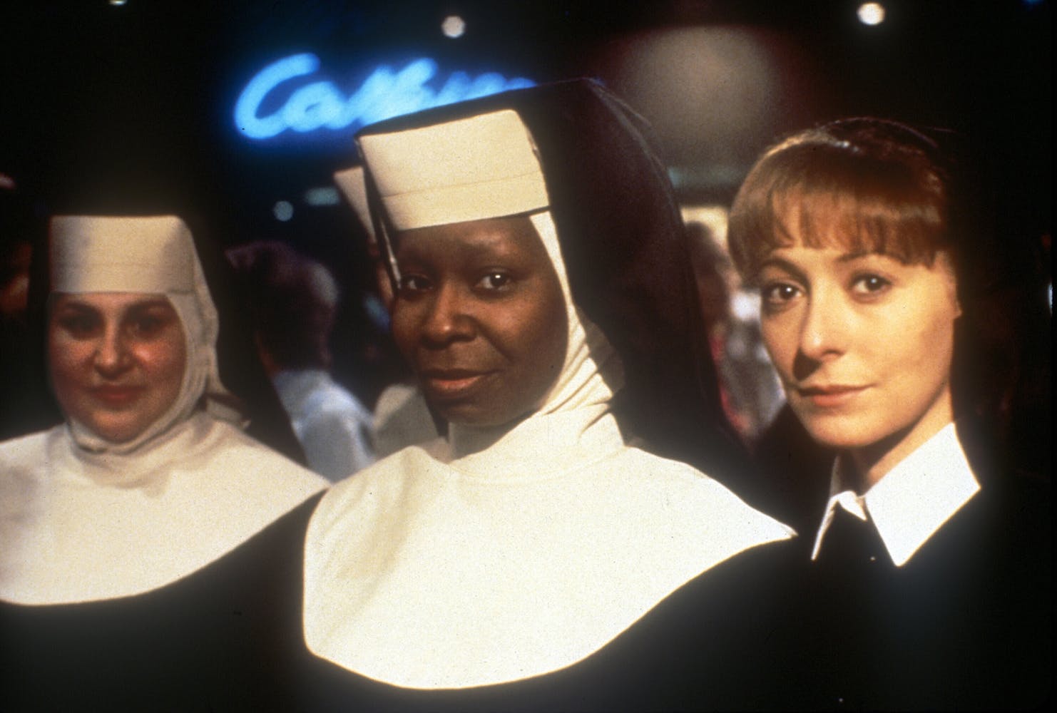 Three women in nun habits look at the viewer while standing in a nightclub.