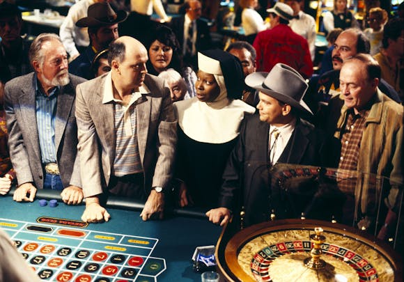 A woman in a nun habits speaks to a group of men in western wear at a craps table in a casino.
