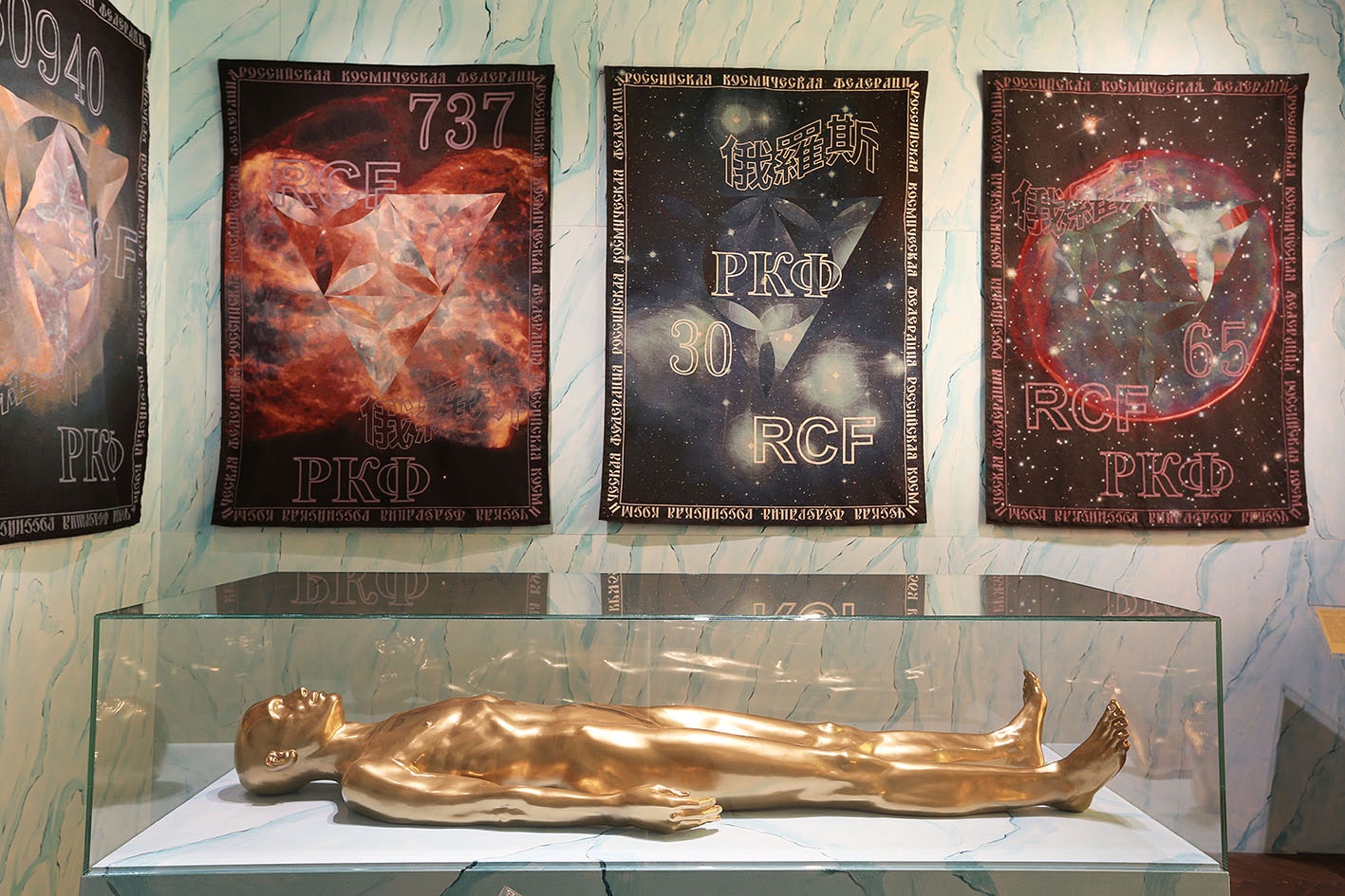 Artwork image of gold human figure within a glass case within an art gallery.