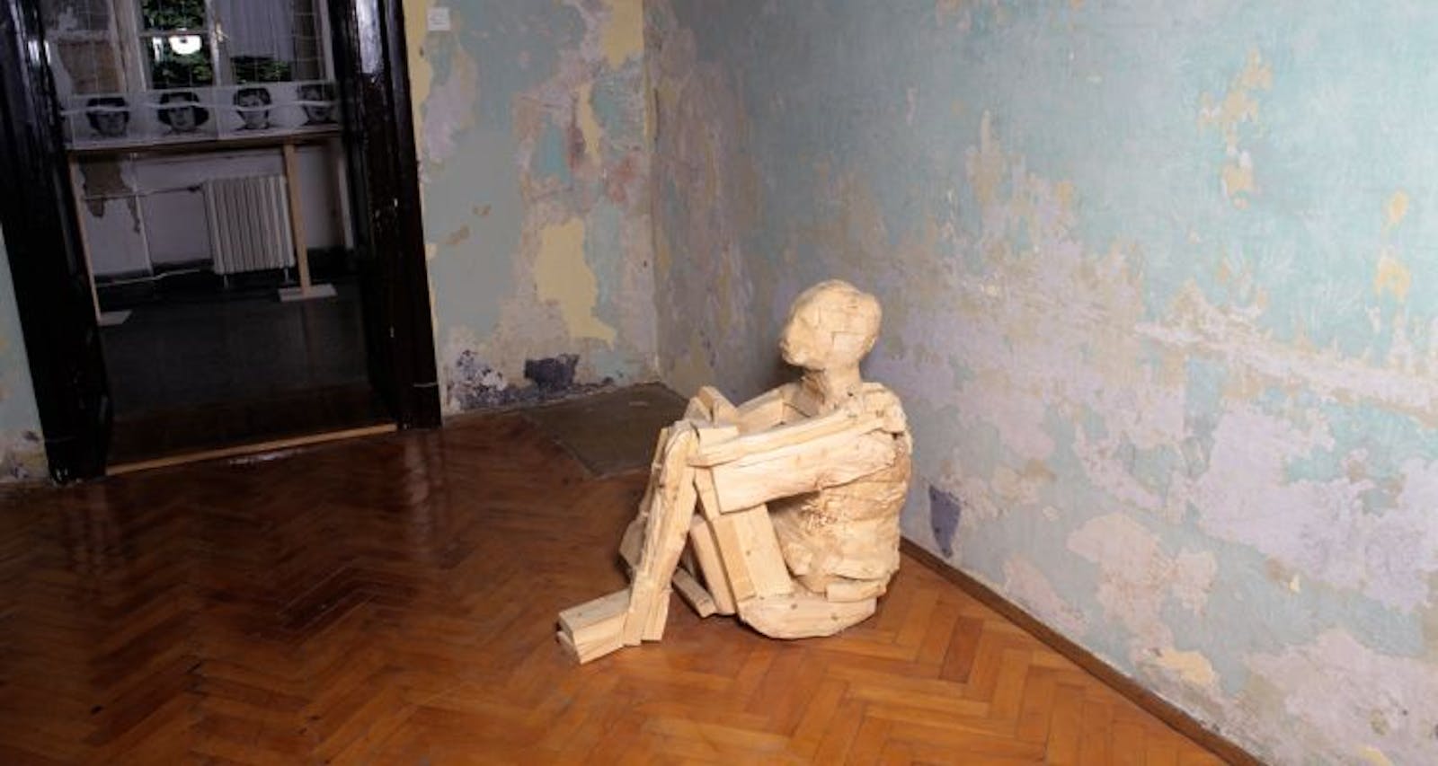 A wooden sculpture of an adult sits in a room with paint peeling walls.