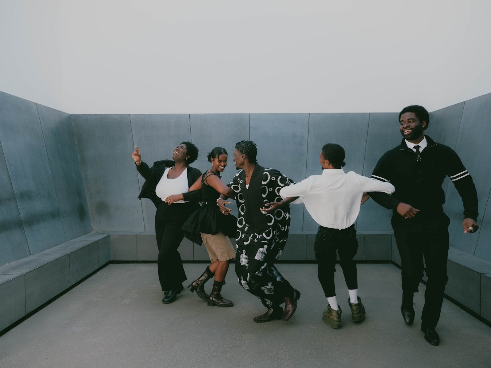 A group of five Black individuals dressed in matching black and white clothes joyfully dance in a minimalist, concrete-walled space.