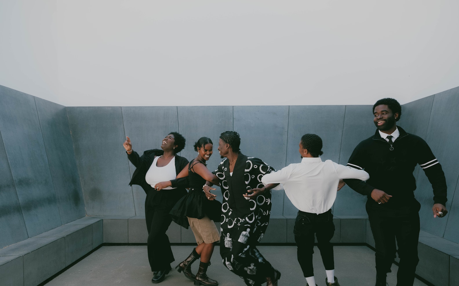 Artropolis_01 A group of five Black individuals dressed in matching black and white clothes joyfully dance in a minimalist, concrete-walled space.