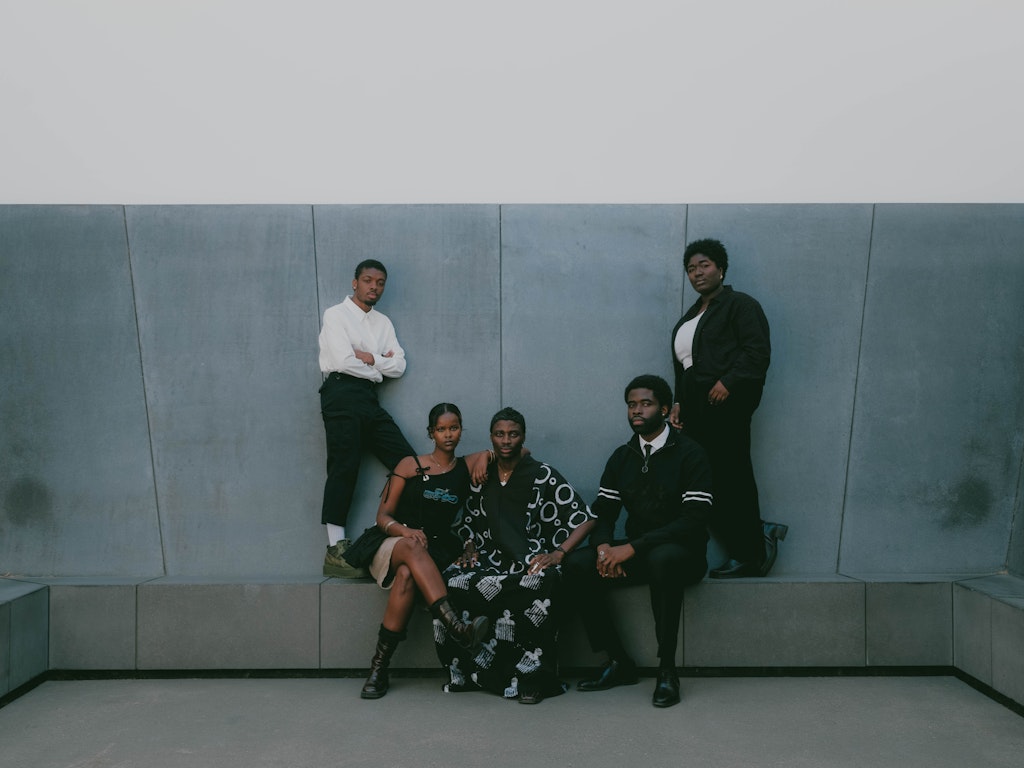 A group of five Black individuals dressed in matching black and white clothes, pose for a wide frame, formal portrait against a gray architectural backdrop. Three are sitting down, and are flanked by two standing people.