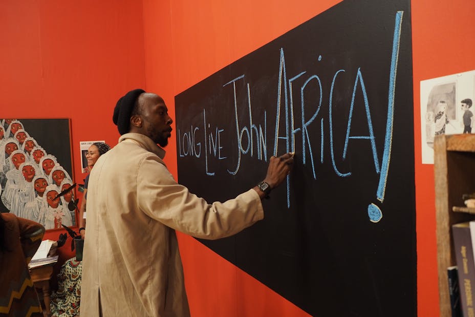 A Black man write on a chalkboard a message that reads Long Live John Africa.