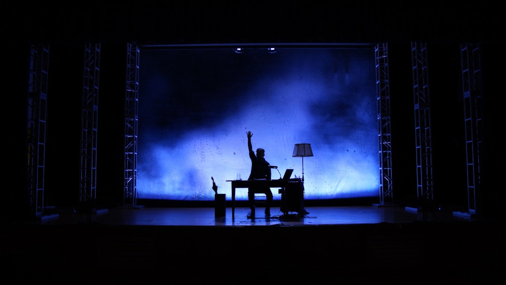 Josh Fox on a stage, in silhouette against a blue background. He is seated at a table with his arm raised in the air.