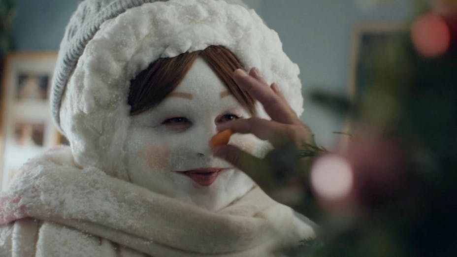 A woman with elaborate makeup to make her look like a snowman. A hand places a carrot on her nose.
