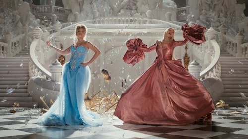 Two women in ball gowns, blue and pink, in a decadent hall with staircases and statues. Bits of shiny things fall around them.