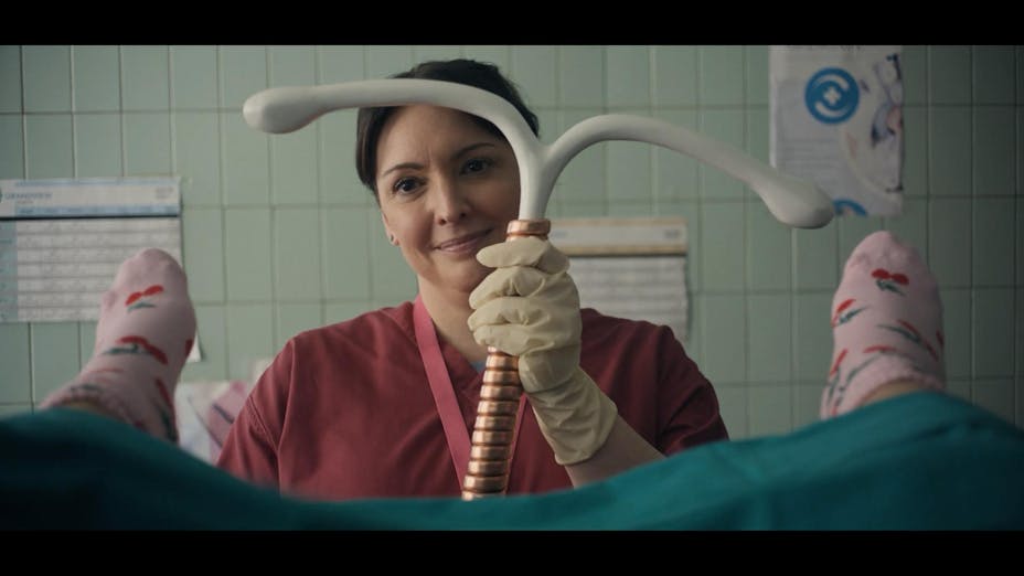 From the point of view of a person on a doctor’s table, we see the edge of a gown and sock feet in stirrups. A nurse smiles holding a gigantic IUD.