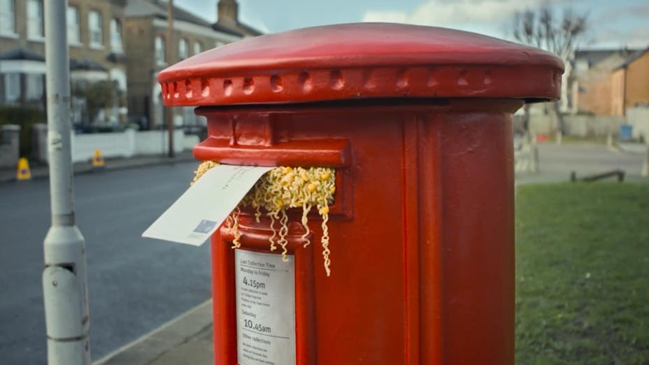 Red British mailbox with ramen noodles stuffed in the slot with an envelope.
