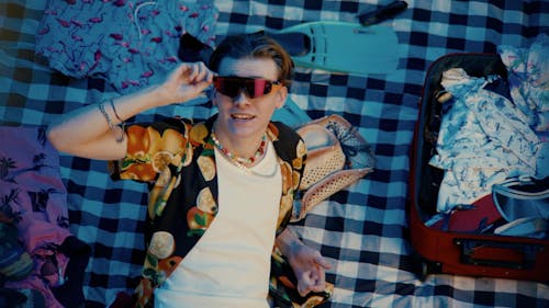 A person with light skin holding sunglasses laying on a gingham fabric with vacation clothing around them.
