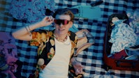 A person with light skin holding sunglasses laying on a gingham fabric with vacation clothing around them.