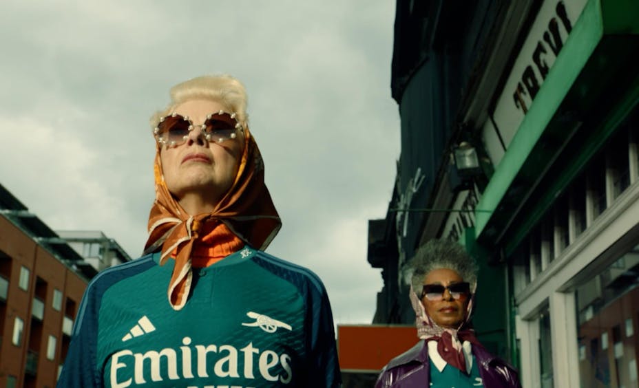 Two women with sunglasses and coiffed hair in silk scarves wearing Arsenal jerseys look confidently ahead down the street.