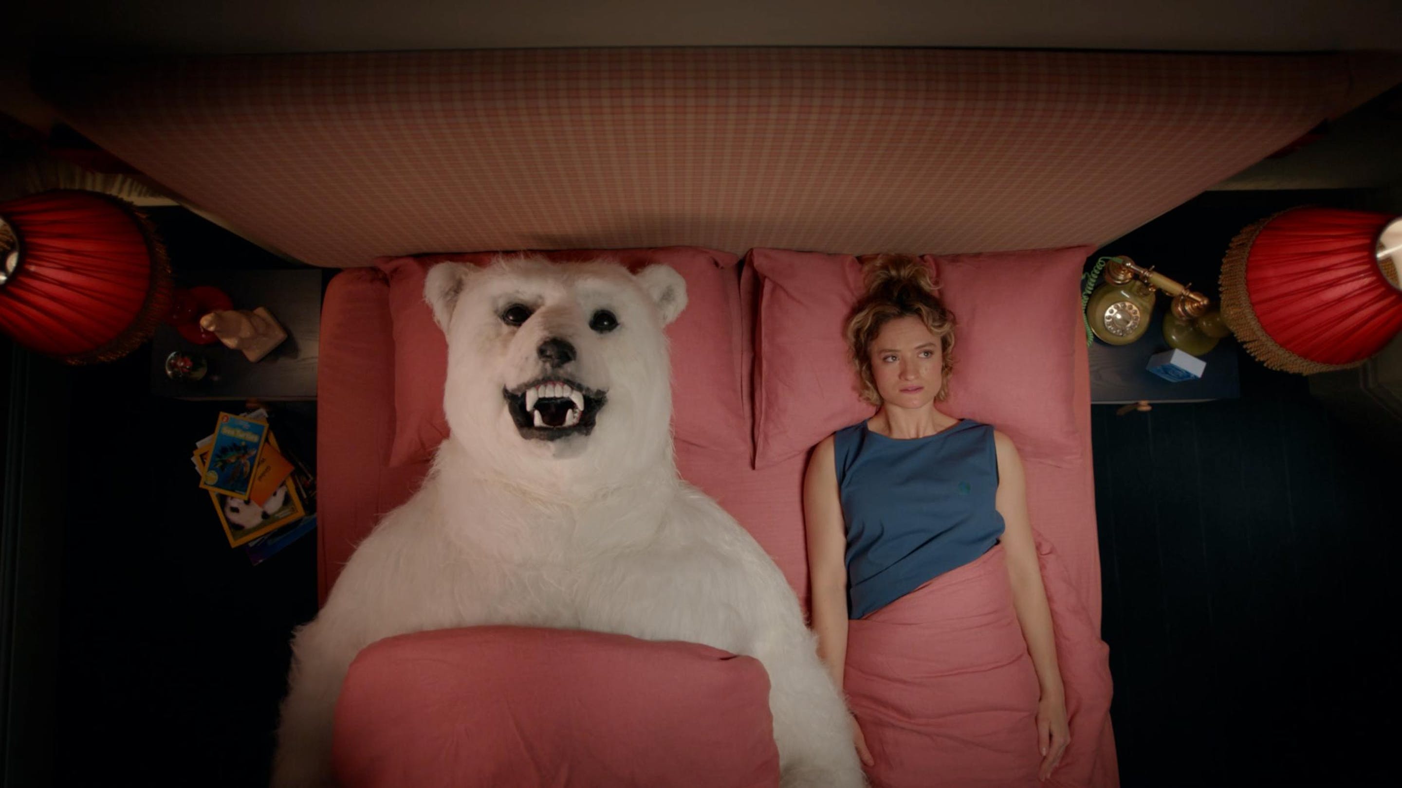 An overhead view of a woman in a blue top laying in a pink bed with a polar bear. They lay side by side on their backs.
