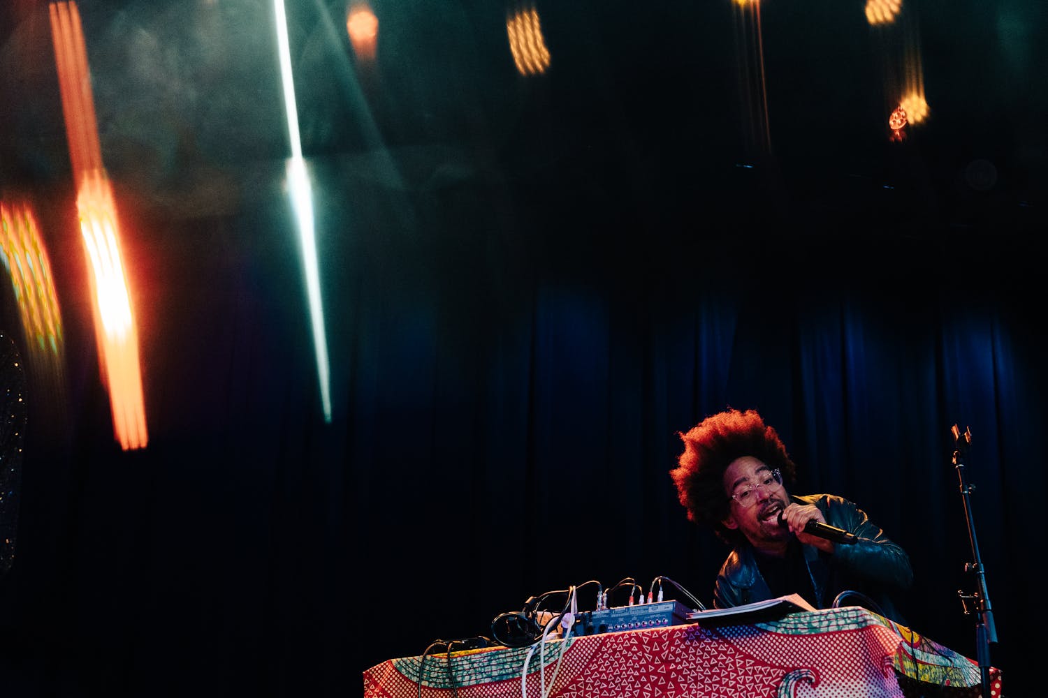 Damon Locks sings into the microphone. He sits behind a table with a red patterned tablecloth and a drum machine.