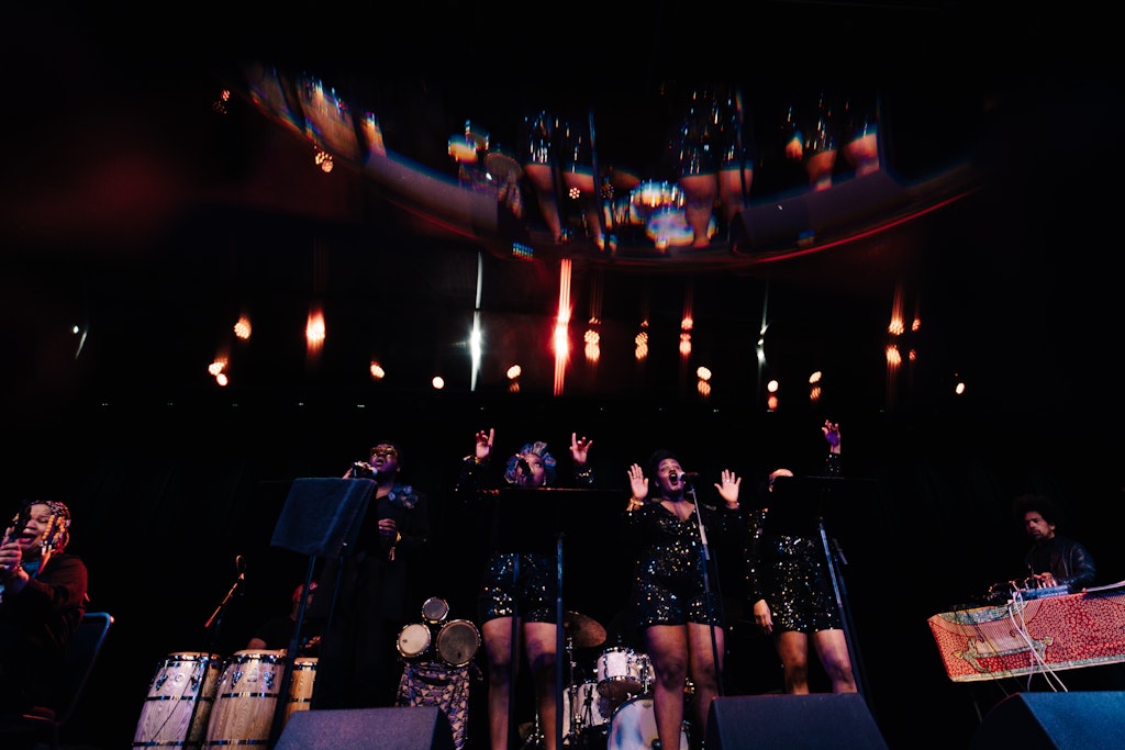 Four Black singers stand center stage during a performance. The three female singers on the right wear short sequined dresses, and the male singer at left wears a black suit. The stage lights make everything shine.