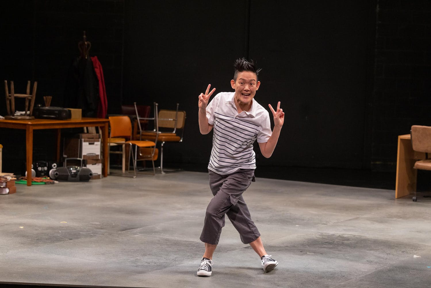 A person with light skin tone and short, spiky hair is wearing a striped polo, grey pants and converse sneakers. They are on stage, giving two peace signs, standing with their legs crossed. A pile of furniture is visible behind them.