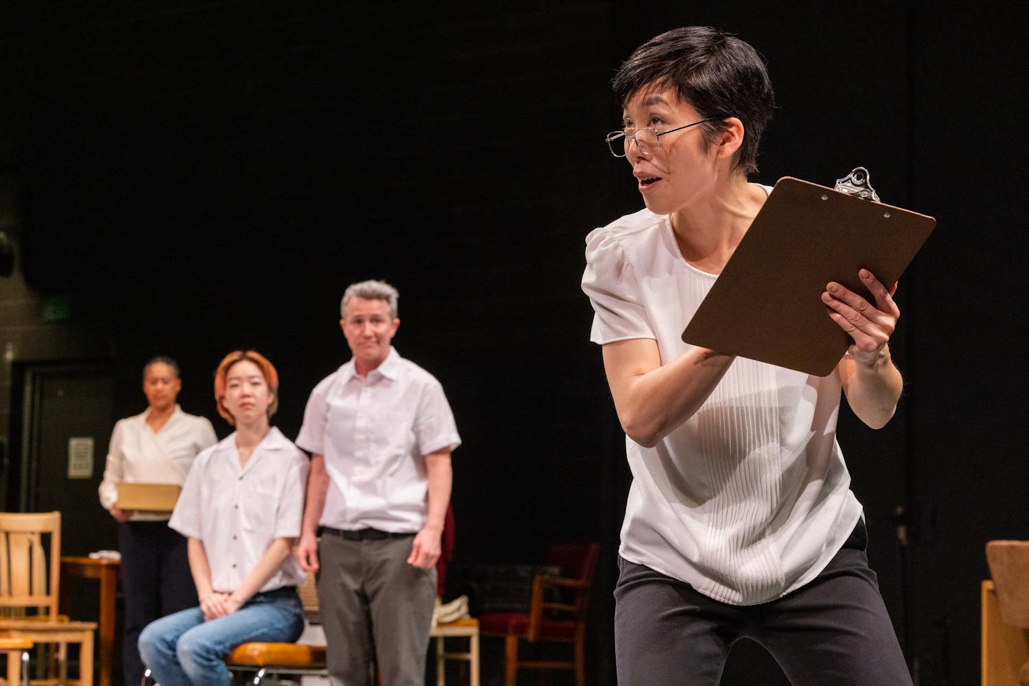 A person with light skin and short, dark hair wears wire glasses low on their nose, carries a clipboard and appears to be talking. Three figures look on in the background.