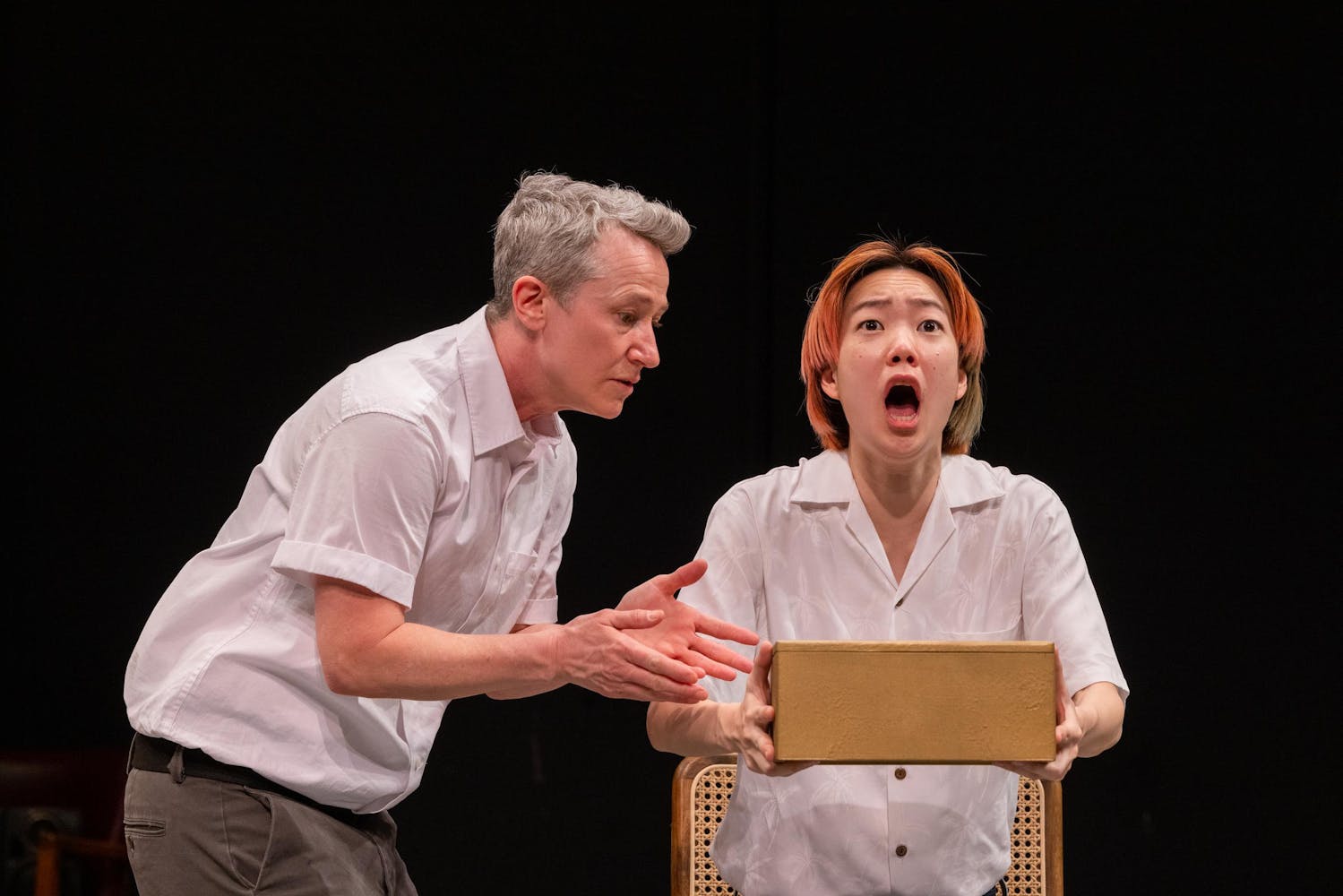 A person with light skin tone and orange hair holds a box in front of them and has their mouth open, expressing perhaps surprise or fear. Another person with light skin and short gray hair is next to them and looks at the box.