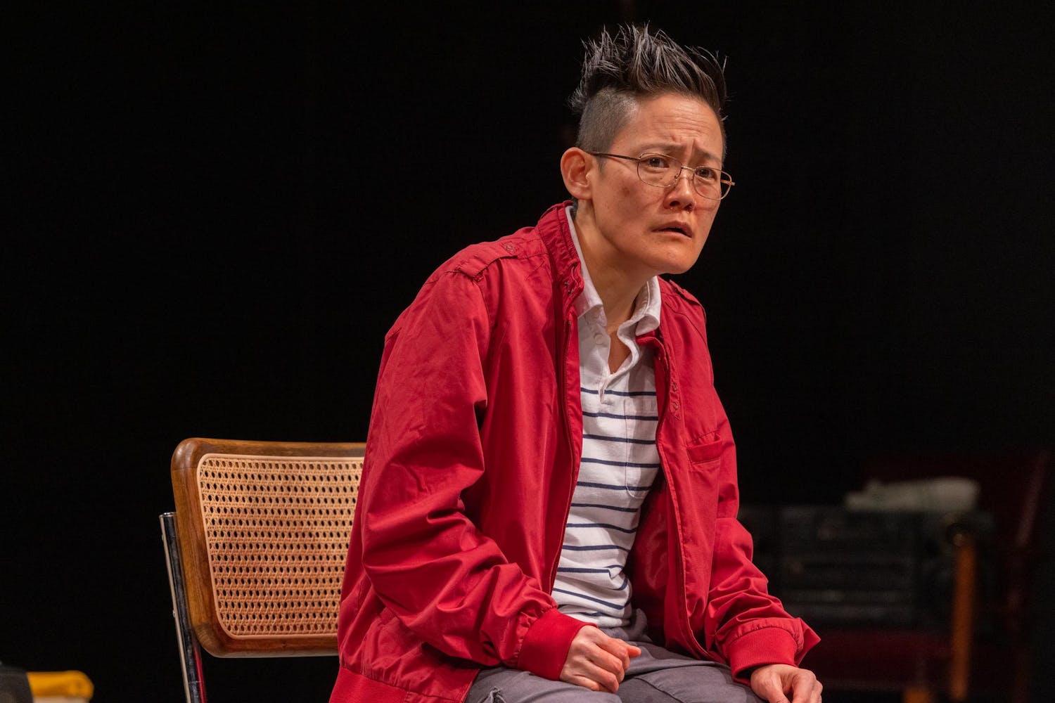 A performer with light/medium skin and short, spiky dark hair sits on a cane back chair on a stage, wearing a red jacket, striped shirt and wire glasses. They look puzzled or concerned.