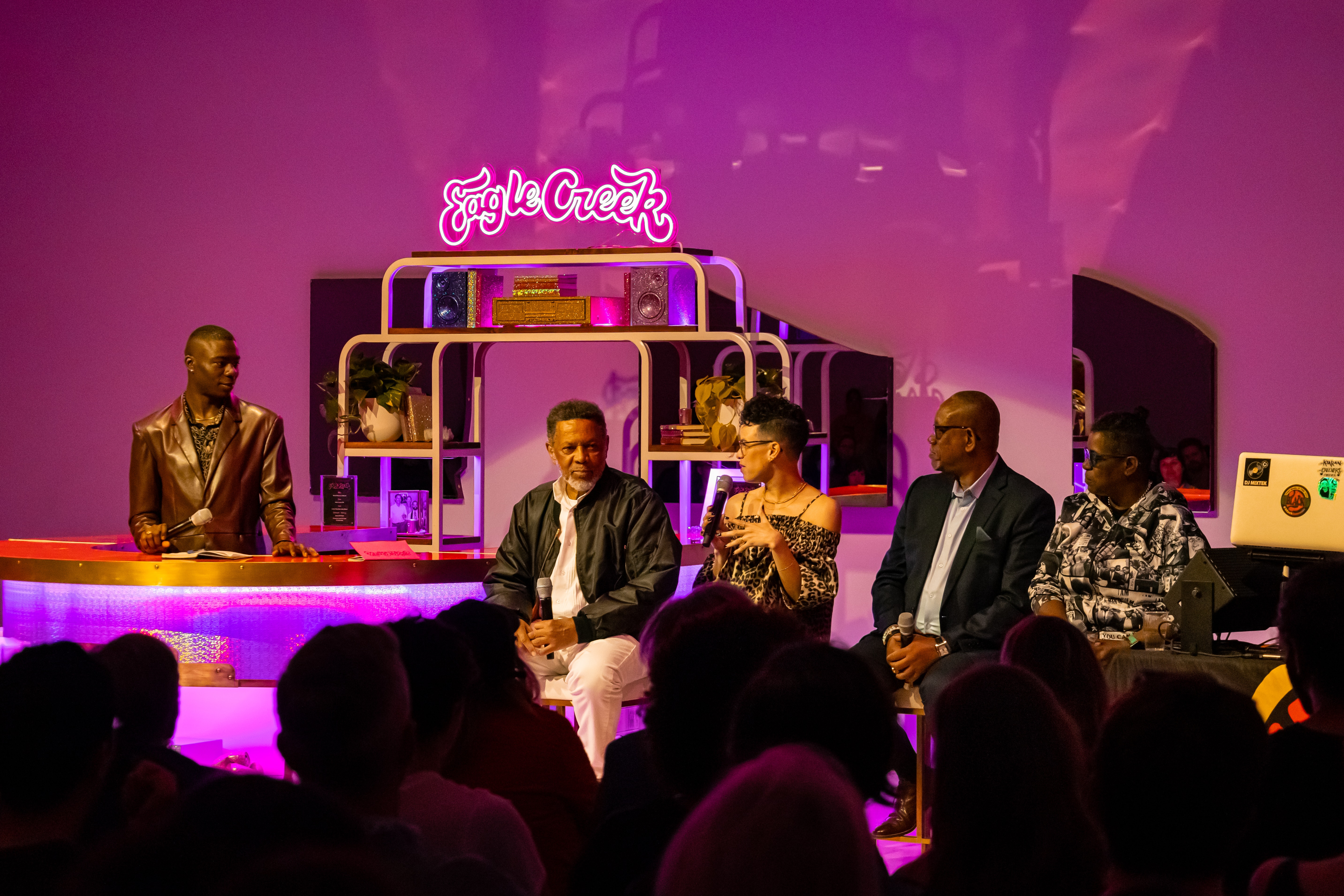 A group of adults with brown skin sit in front of a purple, lit-up bar in a space. Neon light reads 