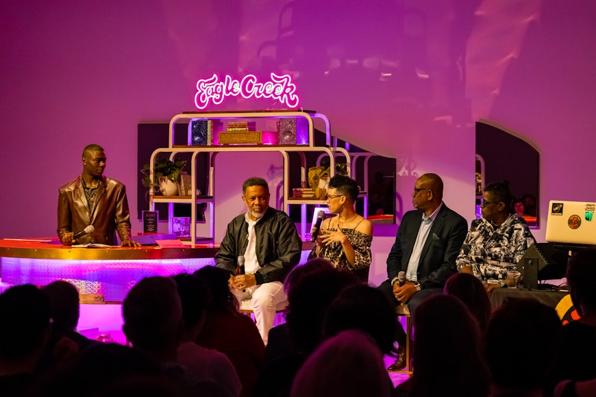 A group of adults with brown skin sit in front of a purple, lit-up bar in a space. Neon light reads "Eagle Creek"