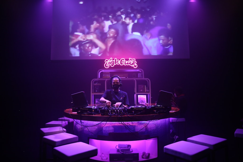 An adult with brown skin stands behind a purple lit bar, playing on mixing boards.