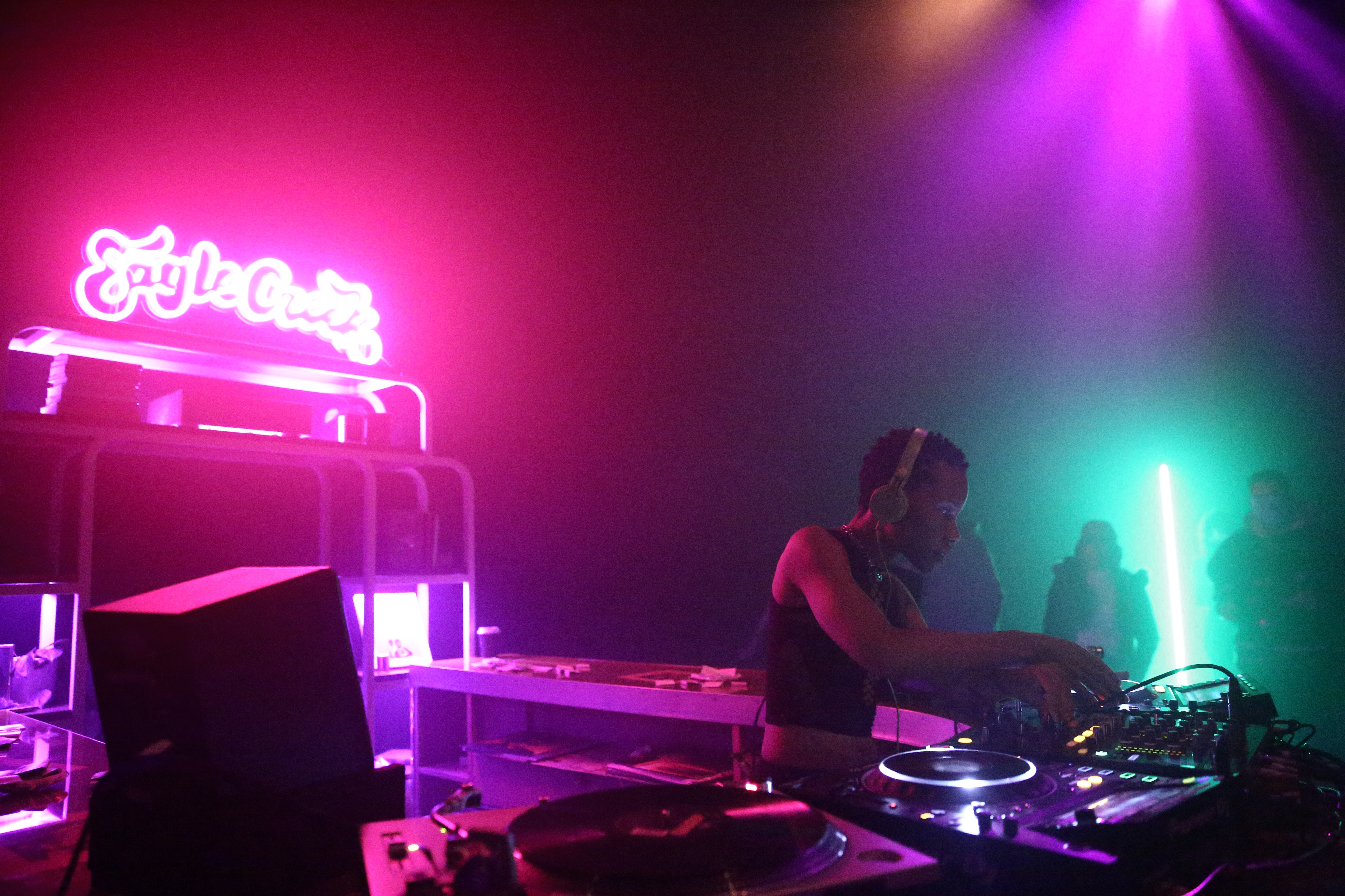 A DJ plays in a colorful lit space. Neon sign reads 