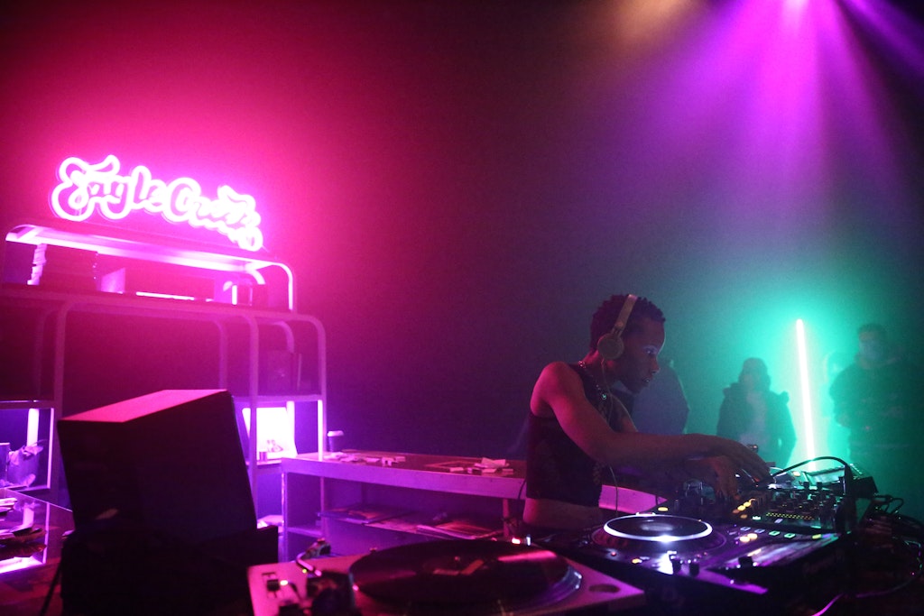 A DJ plays in a colorful lit space. Neon sign reads 