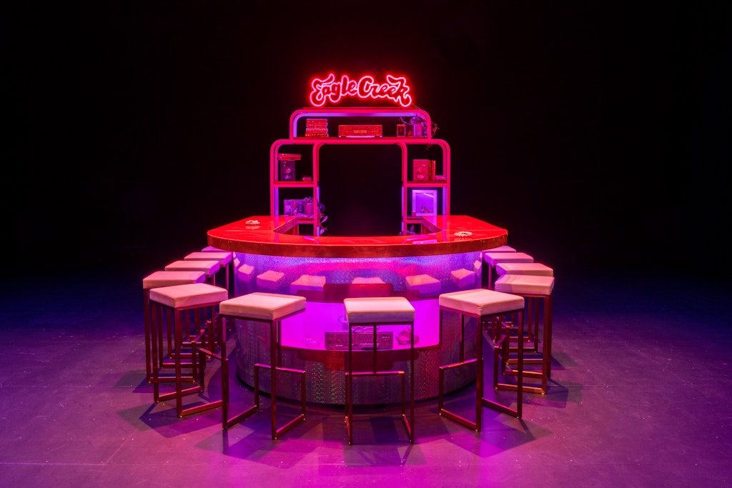 A red and purple-lit bar installation in a black space. Light up sign on top reads 
