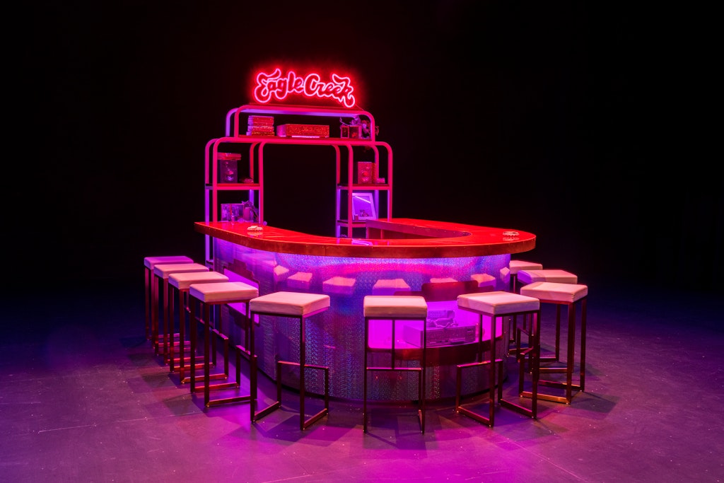 Installation view of Sadie Barnette's The New Eagle Creek Saloon. A restaurant bar with metallic and red details is illuminated by neon pink lights in a dark, empty room. Twelve chairs surround the U-shaped countertop. A neon red sign above the bar reads, 