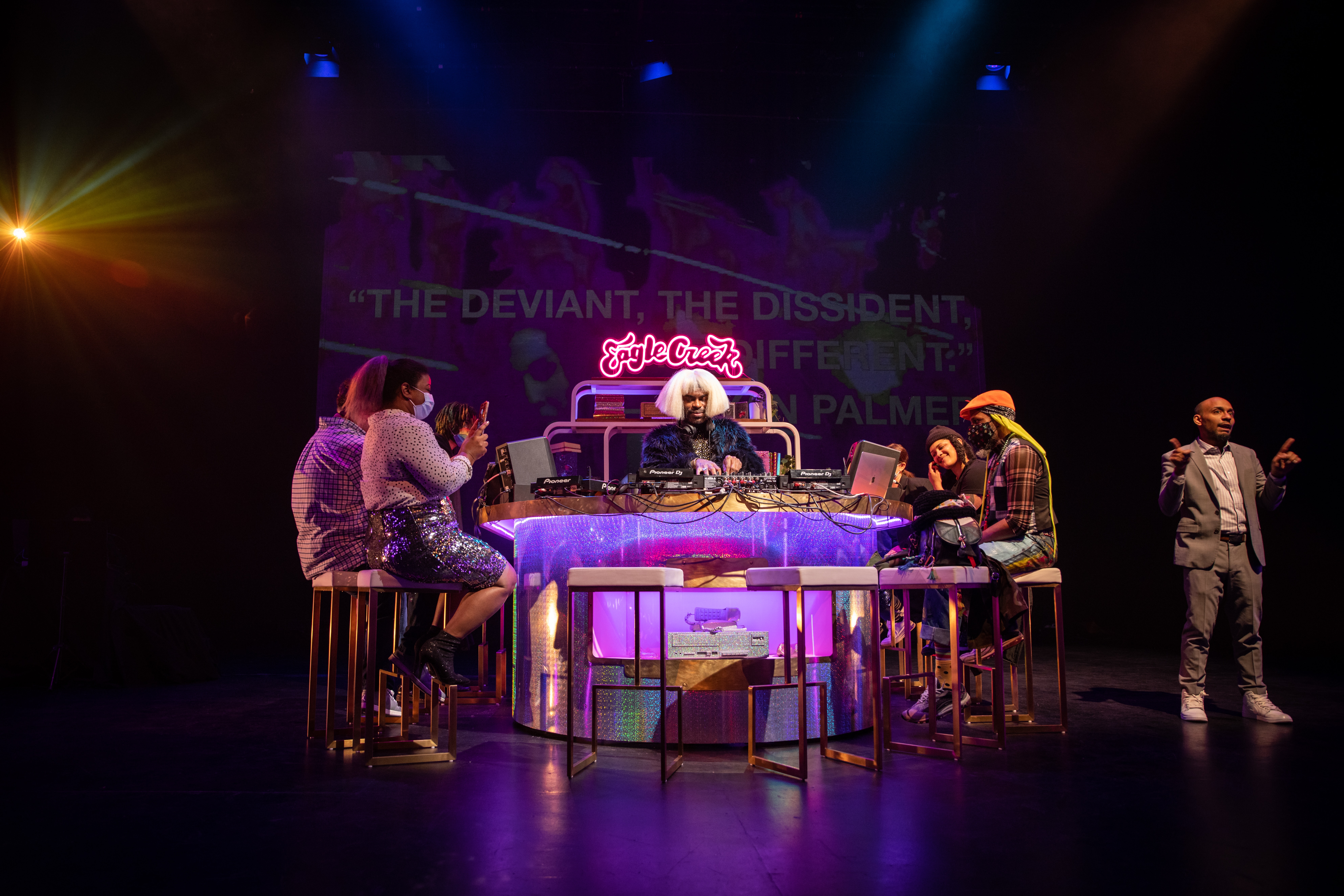 A group of adults sit at a purple lit up bar space. A DJ plays in the center.