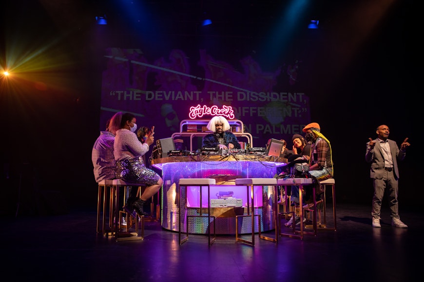 A group of adults sit at a purple lit up bar space. A DJ plays in the center.