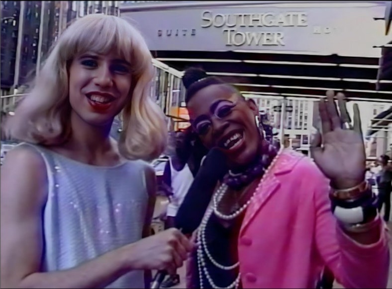Still of a person with light skin and a blonde wig holding a microphone with a person with dark skin in a pink jacket waving.
