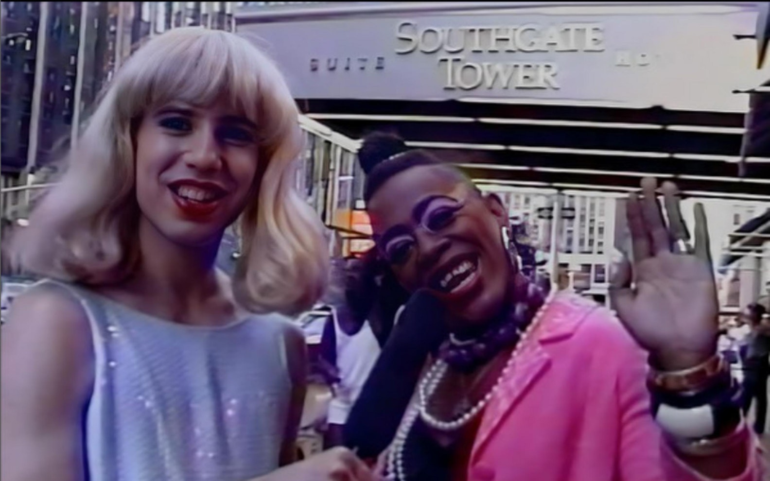 Belverio_On_the_Campaign_Trail_01_1.jpg Still of a person with light skin and a blonde wig holding a microphone with a person with dark skin in a pink jacket waving.