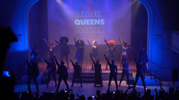 stage of Black Queens Matter event with performance group onstage right arms raised.