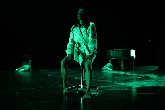 A person sits on a stool on a stage lit with green lights. They have light skin and long dark hair and are wearing a trench coat. Two performers are on the ground in the background, along with a piano.