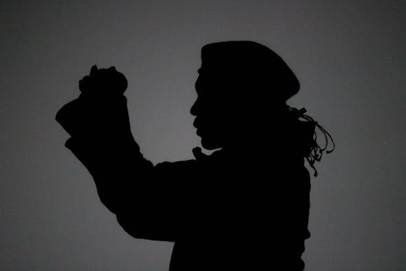In black and white, silhouette, a side view of a figure is shown from the chest up, holding their arms in front of them and wearing a beret.