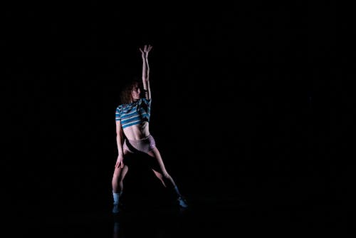 Against an all black background, a person with light skin, dark brown curly hair, a striped crop top and shiny underwear poses with one arm straight up and the other arm resting against their thigh.