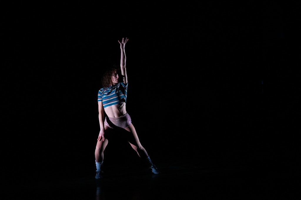 Against an all black background, a person with light skin, dark brown curly hair, a striped crop top and shiny underwear poses with one arm straight up and the other arm resting against their thigh.
