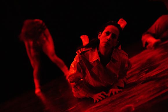 On stage lit with red lights, a dancer with dark brown hair and light skin lays on the ground with their feet lifted behind them, looking at the camera. Two other figures are obscure in the background.