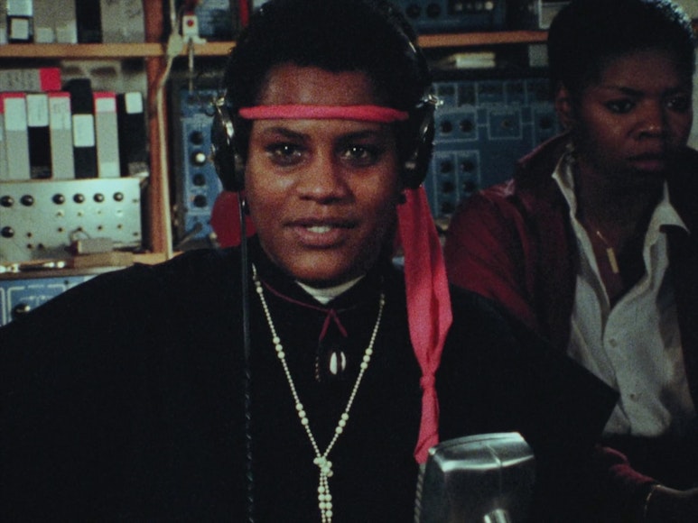 A still of two women. One sits in front of a microphone wearing headphones and a red scarf tied around her forehead.