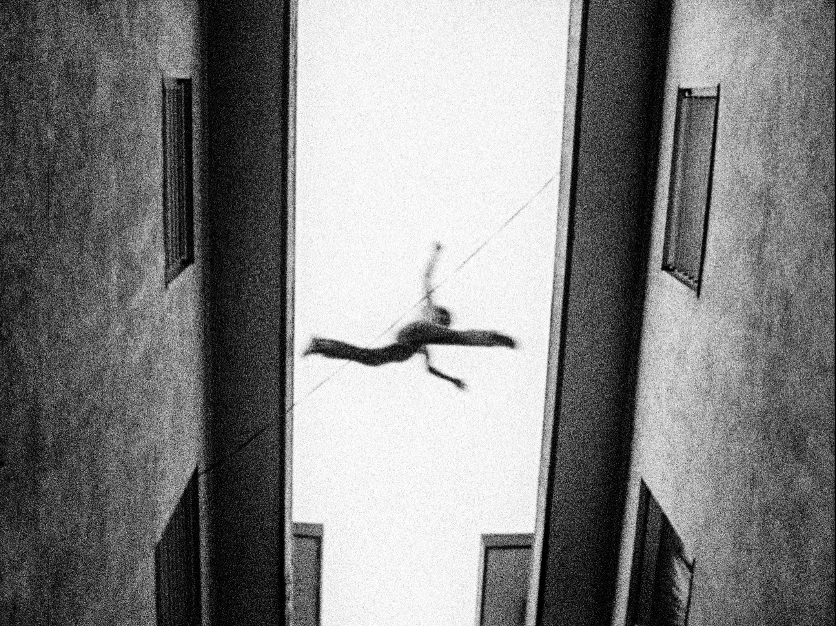 Black and white still shot from below of a young person leaping between the roofs of two buildings. They are mid-jump, not touching either side.