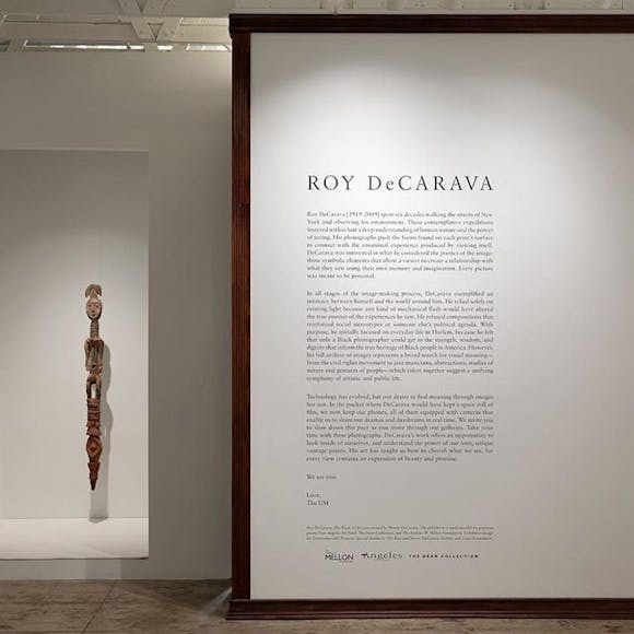 Image of entrance to gallery, with a tall sculpture on the wall and exhibition text in the foreground
