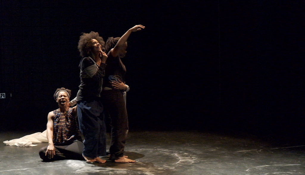 A group of three adult dancers stnad on a black stage and motion away from themselves.