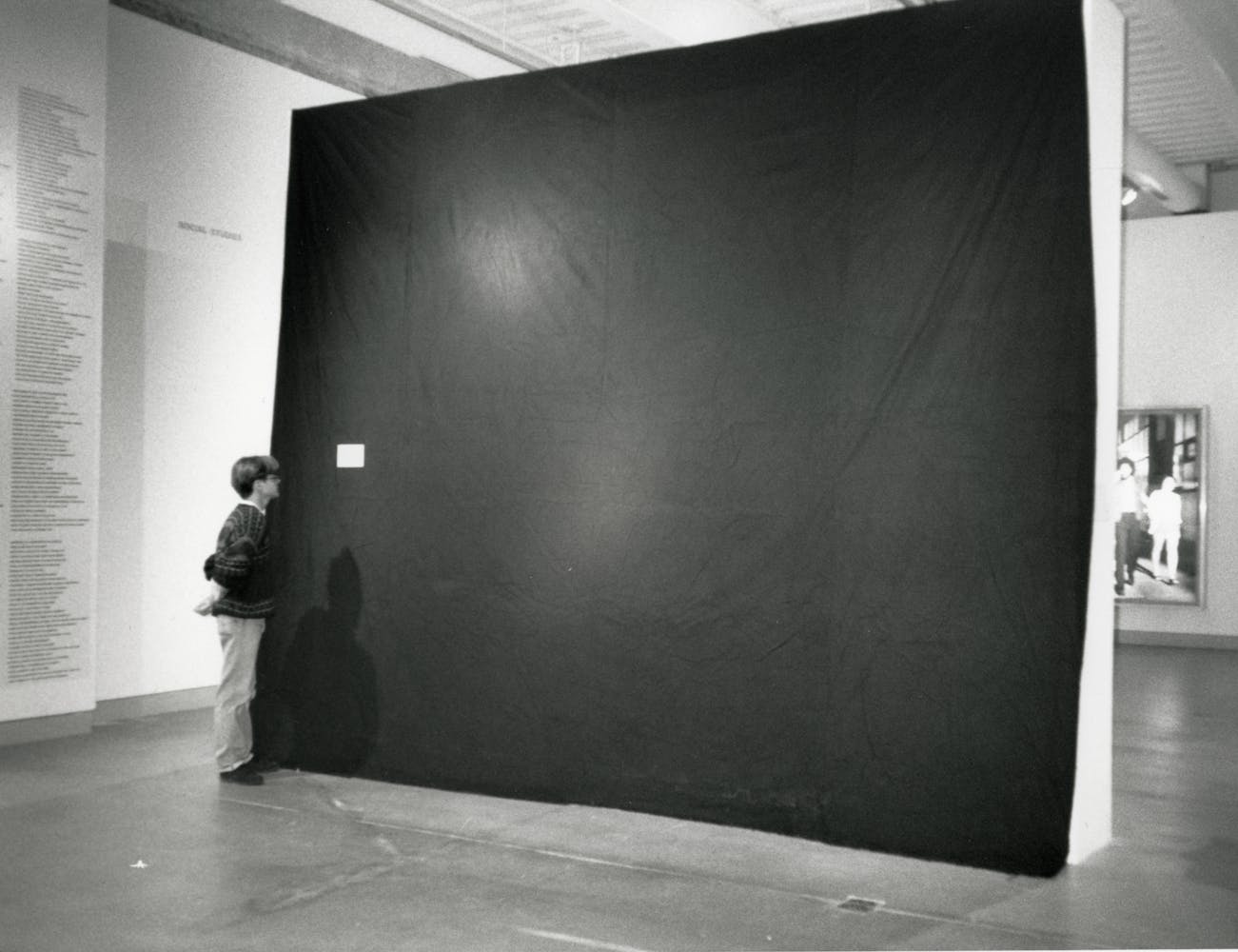 An adult stands close to a black wall in a gallary space, reading the plaque in a black and white photo.