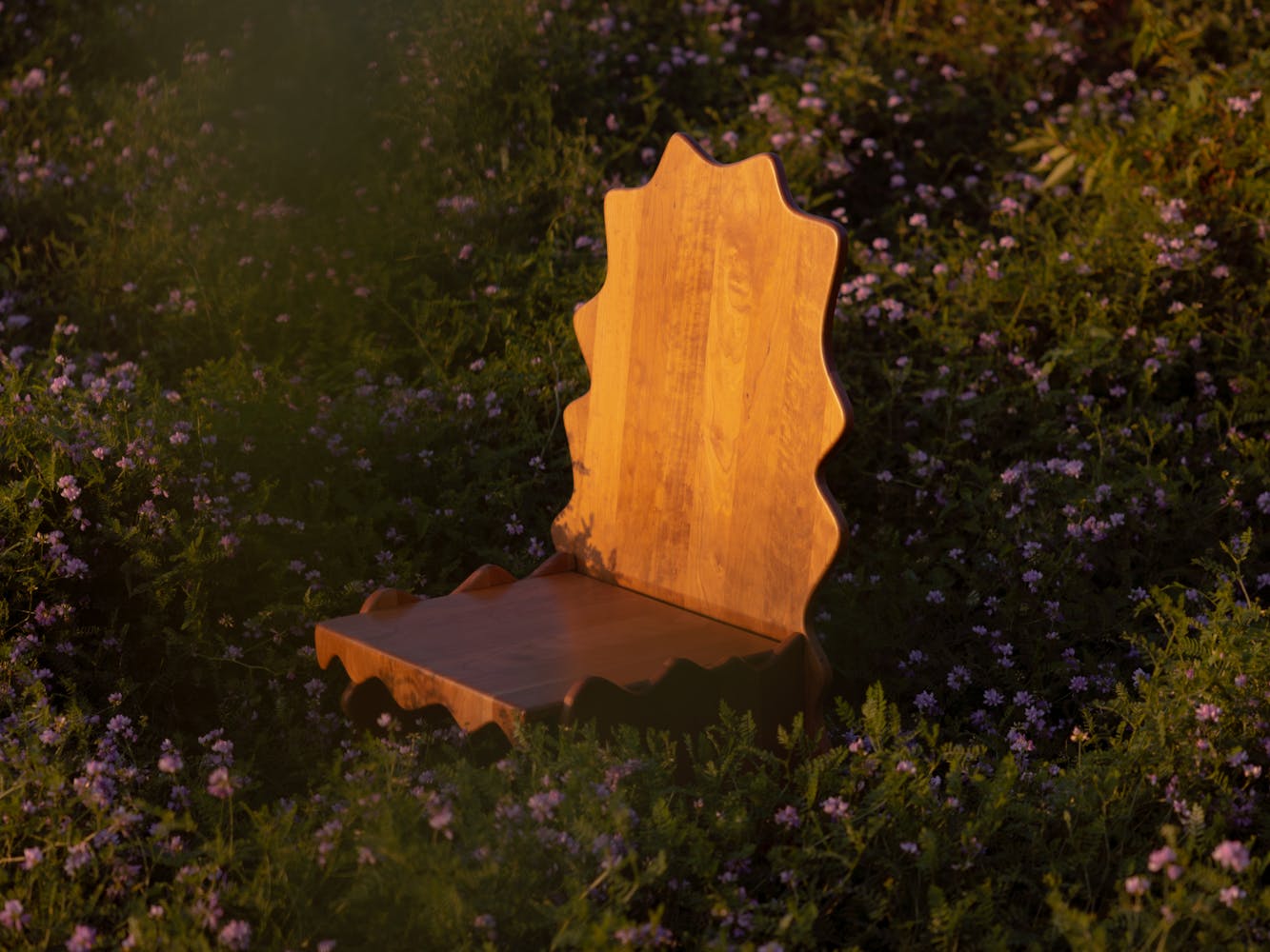 A wavey wooden chair sits amongs flowers outside.