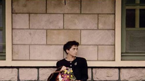In the center of the frame a woman with dark hair and dark clothing holding a bouquet of flowers sits outdoors against a stone building and looks left.
