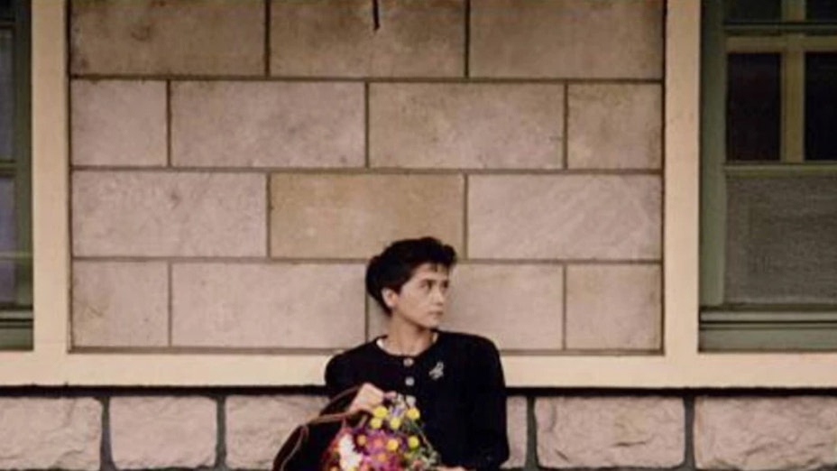 In the center of the frame a woman with dark hair and dark clothing holding a bouquet of flowers sits outdoors against a stone building and looks left.